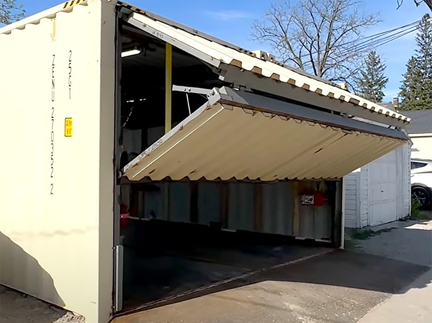 Exterior view of Schweiss bifold shipping container door installed Jeffrey's custom garage shown opening from the side, giving a small peek at the lift straps